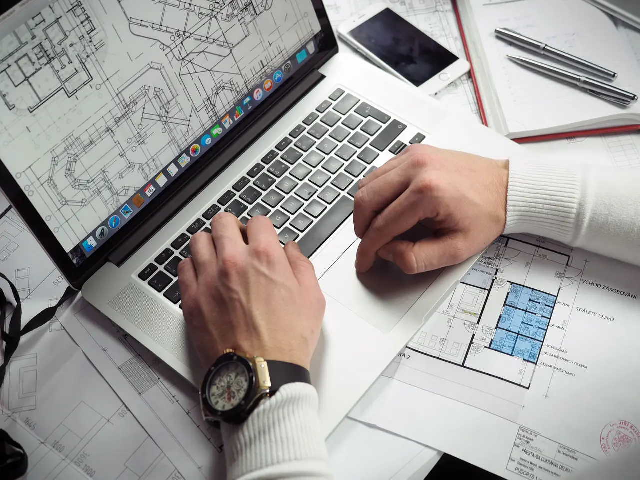Ontwerper die aan een 3D-plattegrond werkt op een laptop, omringd door afgedrukte tekeningen